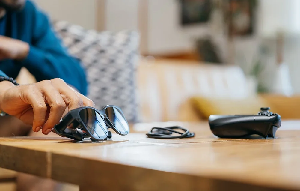 Roka sniedzas pēc melnām XR brillēm, kas guļ uz koka galda blakus kabelim un spēļu kontrolierim