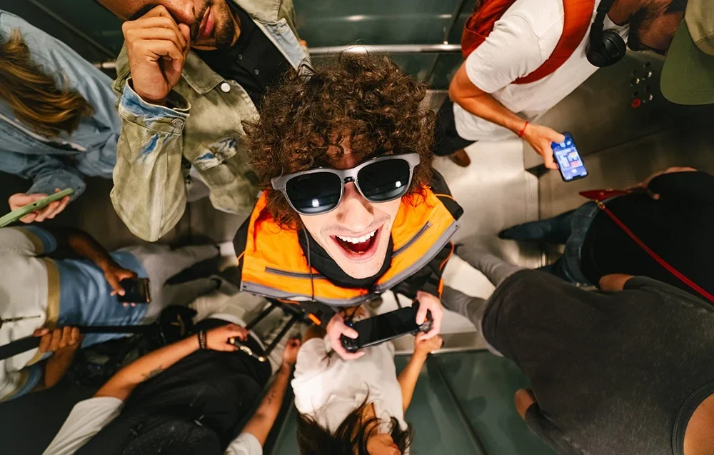 Persona oranžā vestē smejas, valkājot XR brilles, ieskauta citu cilvēku