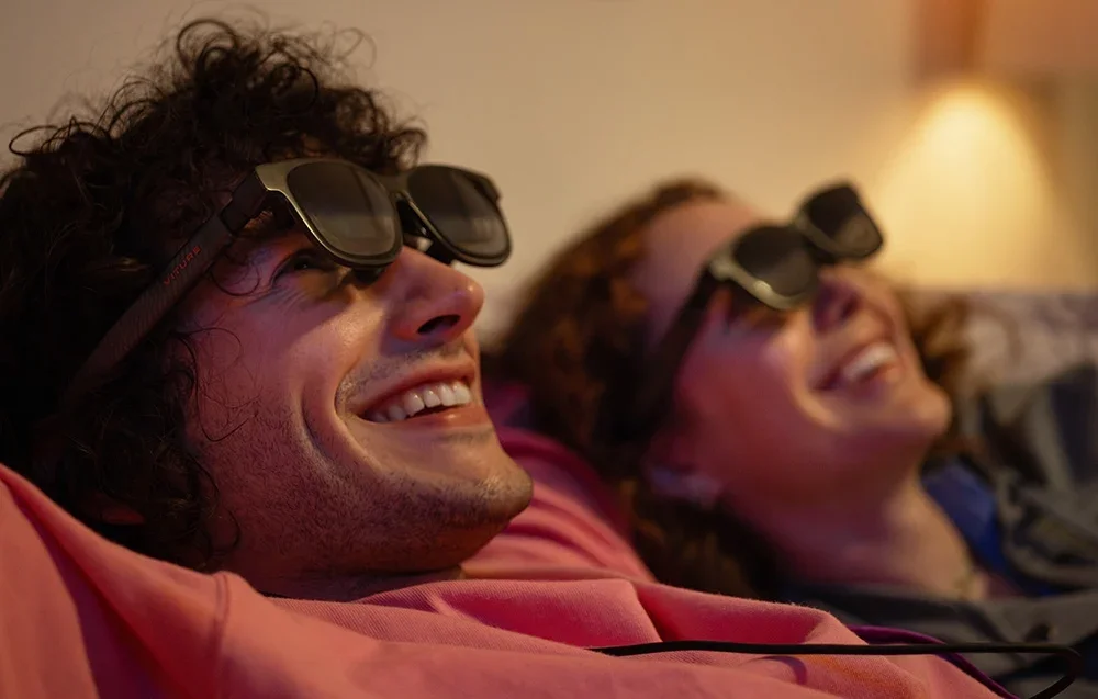 Sieviete un vīrietis ar XR brillēm smejas, guļot istabā ar siltu apgaismojumu