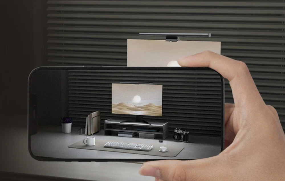 A person holding a smartphone with a view of a computer workstation with a monitor and lamp